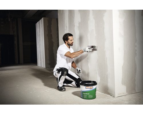 Un homme lisse un mur avec une spatule à côté d''un seau d''enduit Modulan Superfinish.