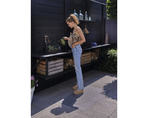Eine Frau gießt mit einer Gießkanne Pflanzen auf einer Terrasse mit Regalen, Holzkisten und einer Bodenmatte.