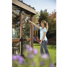 Eine Frau reinigt mit einem Fensterwischer die Scheiben eines Gewächshauses im Garten.