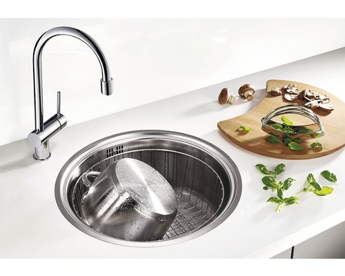 Scène de cuisine avec évier rond, robinet, planche à découper, casserole et ingrédients.