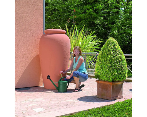 Frau befüllt eine grüne Gießkanne an einem terracottafarbenen Regenwassertank im Garten.