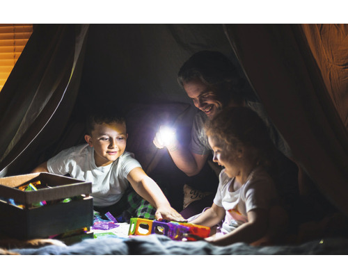Vater und Kinder spielen mit einer Taschenlampe und Spielzeug in einem Zelt.