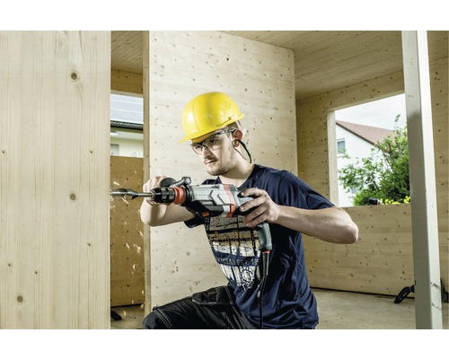 Un homme perce un trou dans une planche de bois avec une perceuse. Il porte un casque de protection et des lunettes de protection.