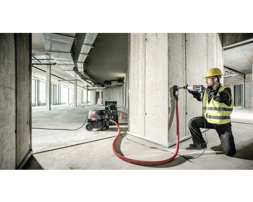 Un ouvrier du bâtiment perce un trou dans un mur en béton avec un marteau perforateur, aspiration de poussière connectée.