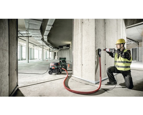 Un ouvrier du bâtiment perce un trou dans un mur de béton avec une perceuse. Un aspirateur industriel et un adaptateur d''aspiration sont également présents.