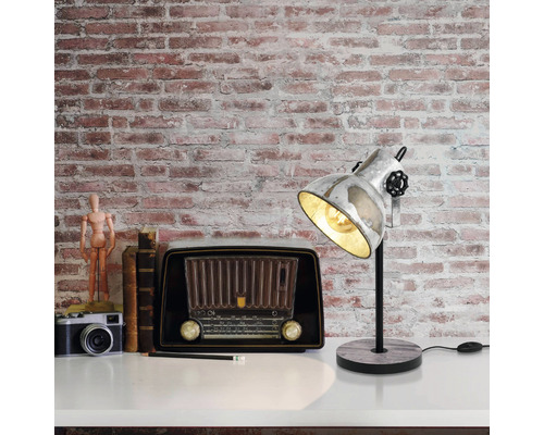 Scène décorative avec une lampe de bureau, une radio, un appareil photo et des livres sur une surface blanche devant un mur de briques.