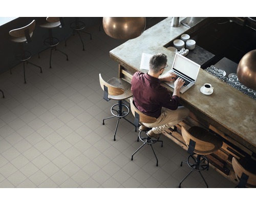 Ein Mann sitzt an einem Tresen auf einem Barstuhl und arbeitet an einem Laptop. Der Raum ist mit quadratischen Bodenfliesen ausgelegt.