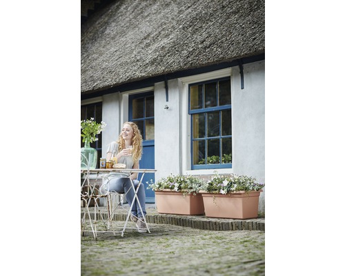 Eine Frau sitzt an einem Tisch vor einem Haus mit Blumenkästen, genießt ihr Frühstück und die entspannte Atmosphäre.