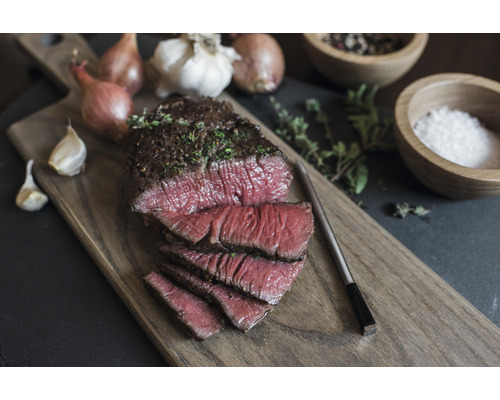 Sur une planche en bois, un morceau de viande tranché, garni d''herbes aromatiques, accompagné d''oignons, d''ail, de poivre et de sel.