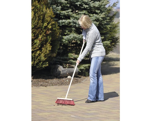 Frau kehrt mit einem Strassenbesen eine gepflasterte Fläche im Garten.