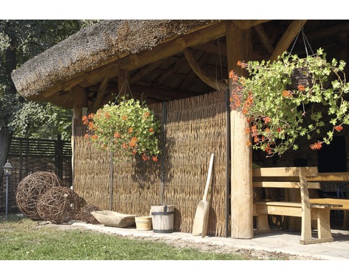 Gartenlaube mit Reetdach, Flechtzaun, hölzerner Sitzgruppe und hängenden Blumenampeln