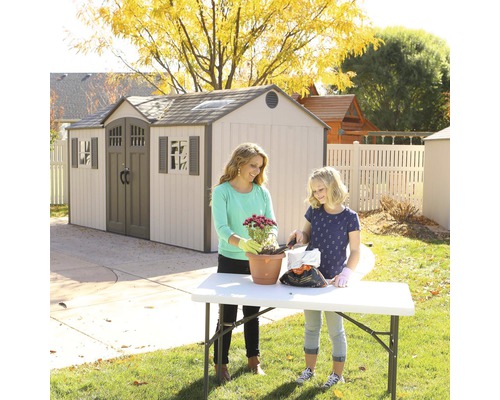Scène avec une remise de jardin en arrière-plan et une femme et une fille plantant des fleurs dans un pot sur une table dans le jardin.