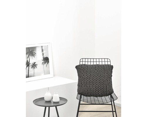 Dekoratives Interieur mit einem grob gestrickten Kissen auf einem Stuhl, einem Beistelltisch und einem gerahmten Bild.