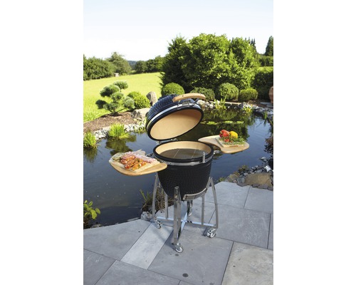 Keramikgrill mit geöffnetem Deckel, Holzablagen und Edelstahlgestell in einer Gartenszene