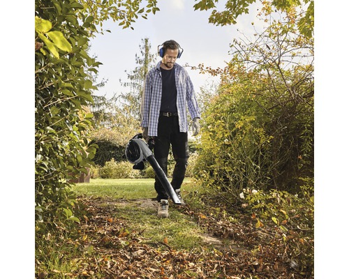 Un homme avec protection auditive et des gants utilise un aspirateur souffleur de feuilles dans le jardin.