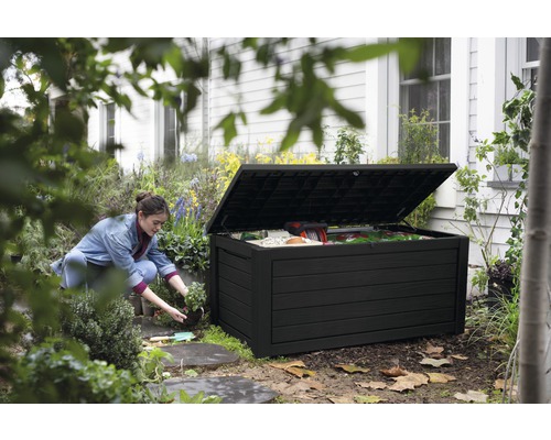 Gartenszene mit einer Frau, die neben einer geöffneten, schwarzen Gartenbox kniet