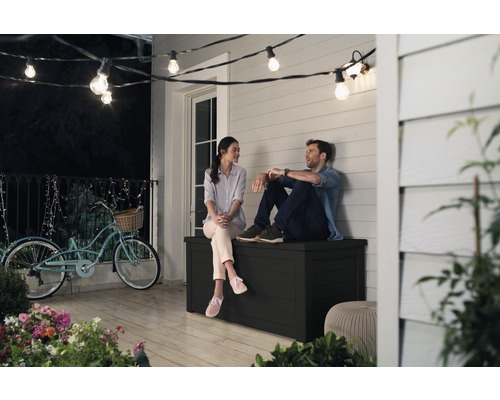 Ein Paar sitzt auf einer schwarzen Gartenbox auf einer Terrasse unter einer Lichterkette.