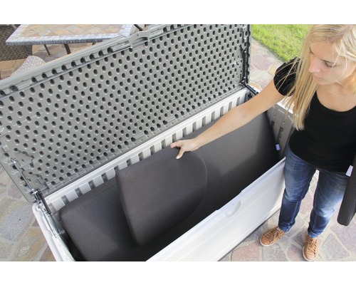 Une femme range des coussins dans un coffre de jardin gris et blanc.