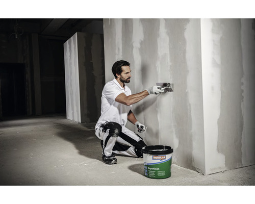 Un homme lisse un mur avec du mastic, tandis qu''un seau de mastic est posé sur le sol.