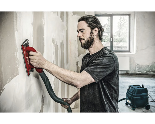 Un artisan ponce un mur avec une ponceuse manuelle raccordée à un aspirateur.