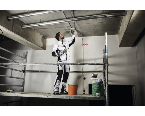 Ein Handwerker auf einem Gerüst verspachtelt eine Decke. Ein Sack Spachtelmasse und ein Eimer stehen auf dem Gerüst.