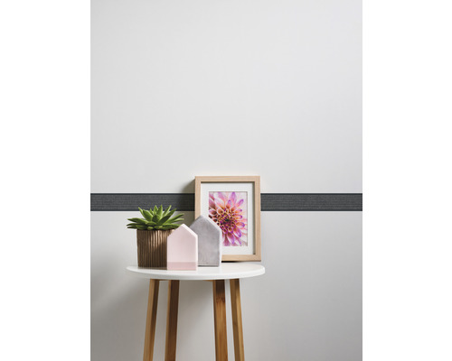 Dekorativer Tisch mit Zimmerpflanze, Dekohäusern und Bilderrahmen vor einer Wand mit Bordüre