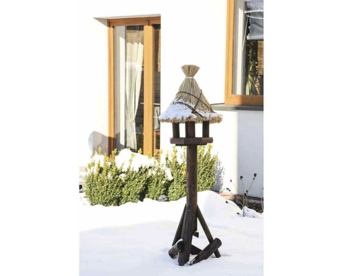 Mangeoire à oiseaux avec toit de chaume sur un support en bois dans un jardin enneigé.
