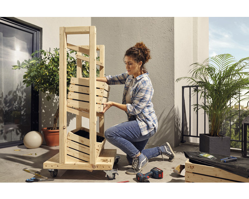 Eine Frau montiert ein Holzregal mit Pflanzkästen auf einem Balkon. Werkzeuge und Pflanzen sind ebenfalls sichtbar.