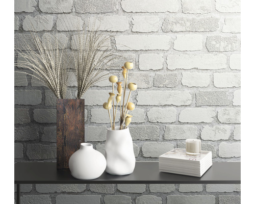 Dekorative Stillleben-Anordnung mit Vasen, Pflanzen und Büchern vor einer Ziegelwand.