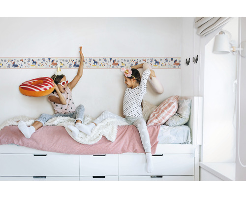 Zwei Mädchen sitzen auf einem Bett mit weißem Rahmen und Schubladen vor einer Wandbordüre mit Einhornmotiv