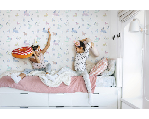 Zwei Mädchen spielen auf einem Bett vor einer Tapete mit Einhornmuster.