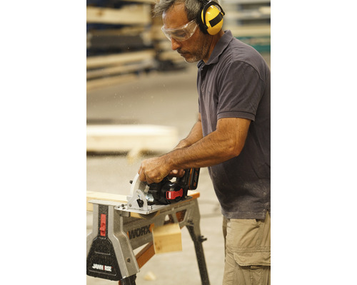 Un homme scie une planche de bois avec une scie circulaire sur une table de travail, portant des lunettes de protection et un casque antibruit.