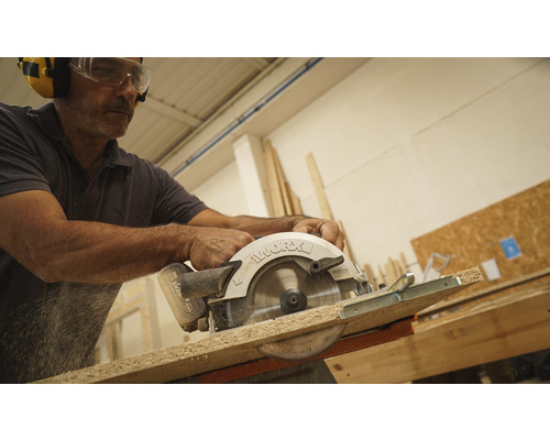 Un homme scie une planche de bois avec une scie circulaire à main, portant des lunettes de protection et des protections auditives.