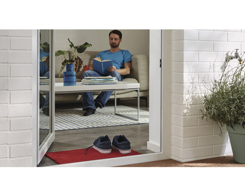 Un homme lit un livre dans un salon, visible à travers une porte ouverte avec un paillasson et des chaussures.