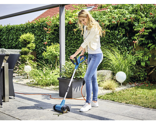 Frau verwendet eine Fugenbürste auf einer Terrasse, um Unkraut zu entfernen.
