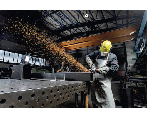 Ein Arbeiter mit Schutzkleidung schleift Metall mit einem Winkelschleifer in einer Werkstatt.
