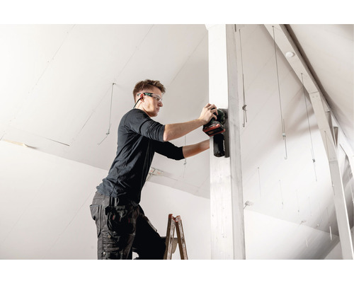 Handwerker auf Leiter beim Schleifen eines Holzpfostens mit einem elektrischen Schleifgerät in einem Dachgeschoss.
