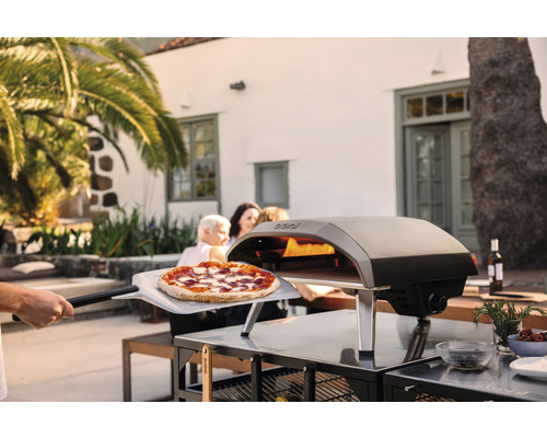 Ooni Pizzaofen im Aussenbereich mit Pizza auf einem Pizzaschieber, umgeben von Gartenmöbeln und Gästen