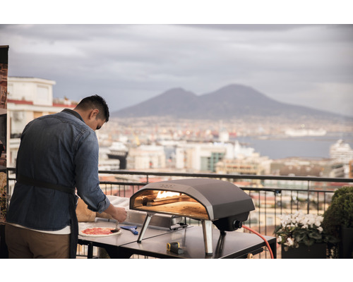 Ein Mann bereitet eine Pizza neben einem Pizzaofen auf einem Tisch im Freien zu.