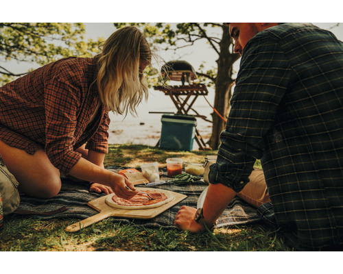 Un couple prépare des pizzas à l''extérieur avec des ingrédients, une pelle à pizza et un four à pizza portable.