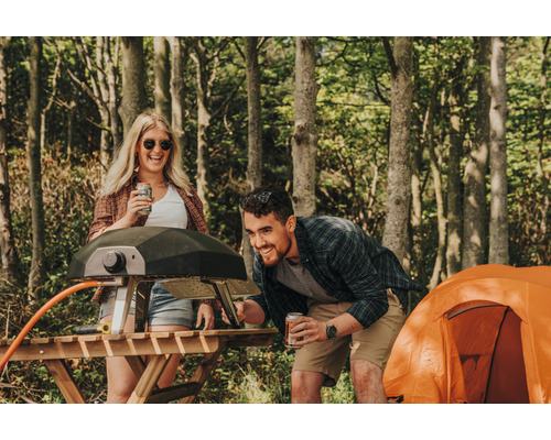 Scène d''extérieur avec une femme et un homme debout à côté d''un barbecue à gaz sur une table en bois, avec une tente en arrière-plan.