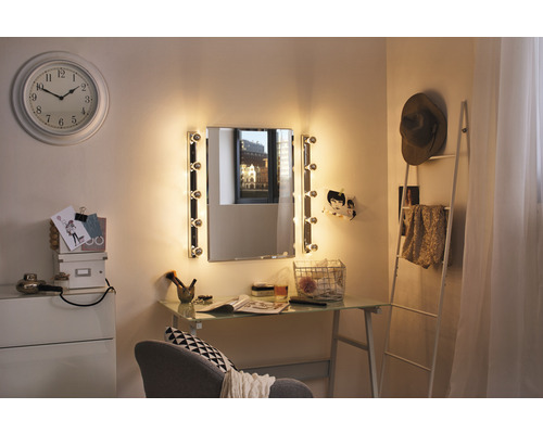 Einrichtung eines hellen Schminktisches mit beleuchtetem Spiegel, Uhr, Kommode und Leiterregal in einem Zimmer.