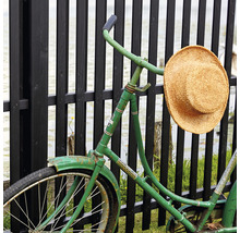 Ein grünes Fahrrad mit einem Strohhut lehnt an einem Zaun.