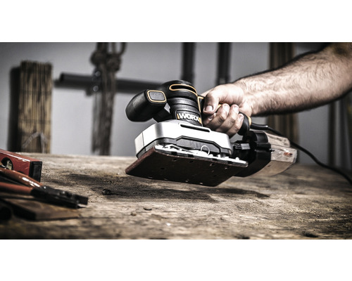 Eine Hand hält einen Schwingschleifer der Marke Worx auf einem Holztisch.