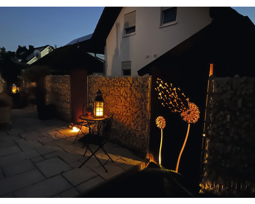Abendliche Gartenszene mit Gabionenwand, dekorativer Metalleinfassung mit Löwenzahnmotiv und Laternenbeleuchtung.