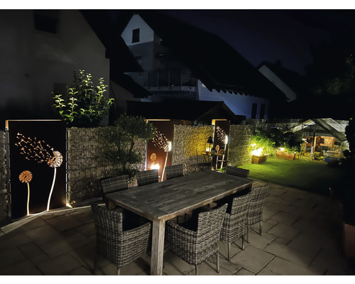 Beleuchteter Garten mit Gabionenzaun, Metallpaneelen mit Löwenzahnmotiven und Esstisch mit Stühlen