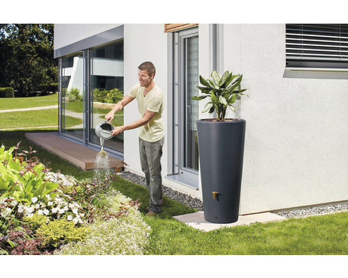 Mann gießt Pflanzen im Garten mit einer Gießkanne, im Hintergrund ein Regenwassertank
