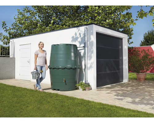 Une femme se tient avec un arrosoir à côté d''une cuve de récupération d''eau de pluie devant un garage.