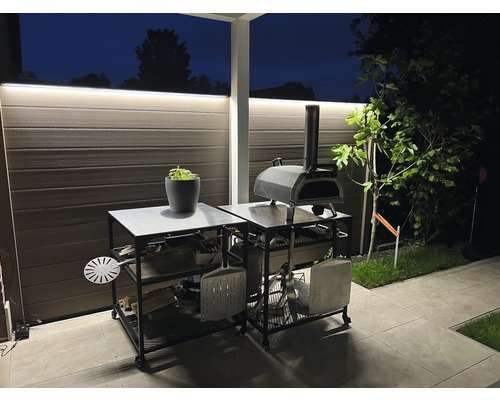 Aussenbereich mit Pizzaofen auf einem Servierwagen mit Regalen für Holz und Zubehör.