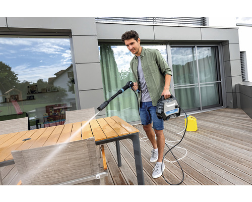 Mann reinigt einen Gartentisch mit einem Hochdruckreiniger auf einer Holzterrasse.